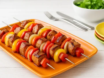 Schaschlik nach DDR-Rezept auf orangefarbener Servierplatte, Spieße mit Fleisch, Paprika, Zwiebeln, Tomaten und Speck, auf weißem Holztisch, dekoriert mit Salatschüssel, modernen Tellern und Besteck.